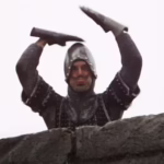 A screenshot from Monty Python & the Holy Grail, showing a knight tapping the top of his head with both hands and sticking out his tongue