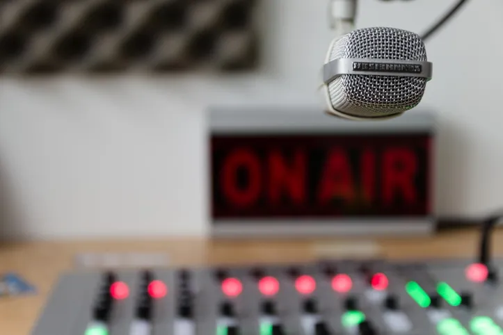 Photo of a studio with an "On Air" sign in the background, and a mixer and microphone in the foreground.
