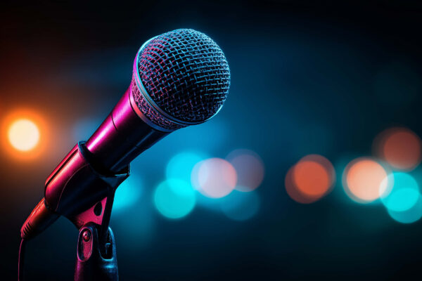 A microphone on a mic stand in the foreground, with blurred colored lights behind it.